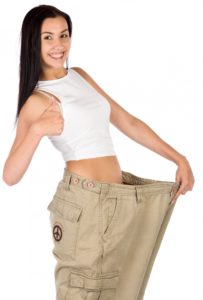brunette woman in white tank top smiling and giving a thumbs up while holding pants that no longer fit to show weight loss